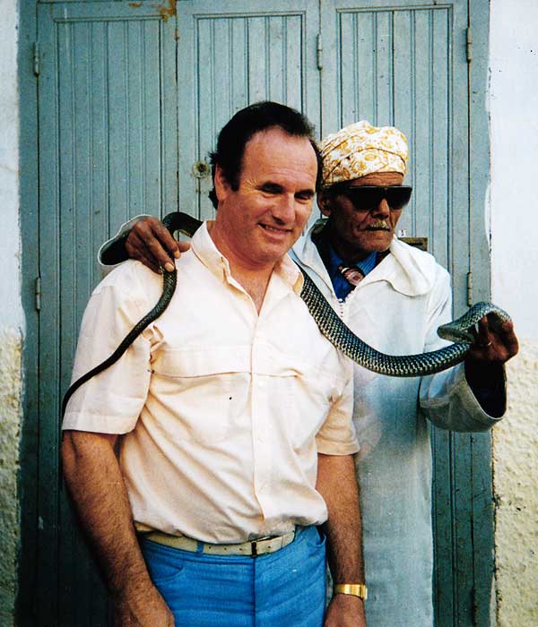 nineteen seventies photo close up of sean in beige shirt and blue trousers in morocco with a snake charmer and his snake wrapped uncomfortably around seans neck