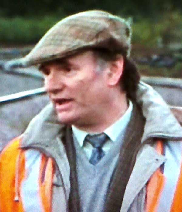 close up front view of sean glenn with head slightly tilted on a jaunty angle dressed in a working mans check fawn coloured suit with v neck grey jumper and tie with a tartan flat cap